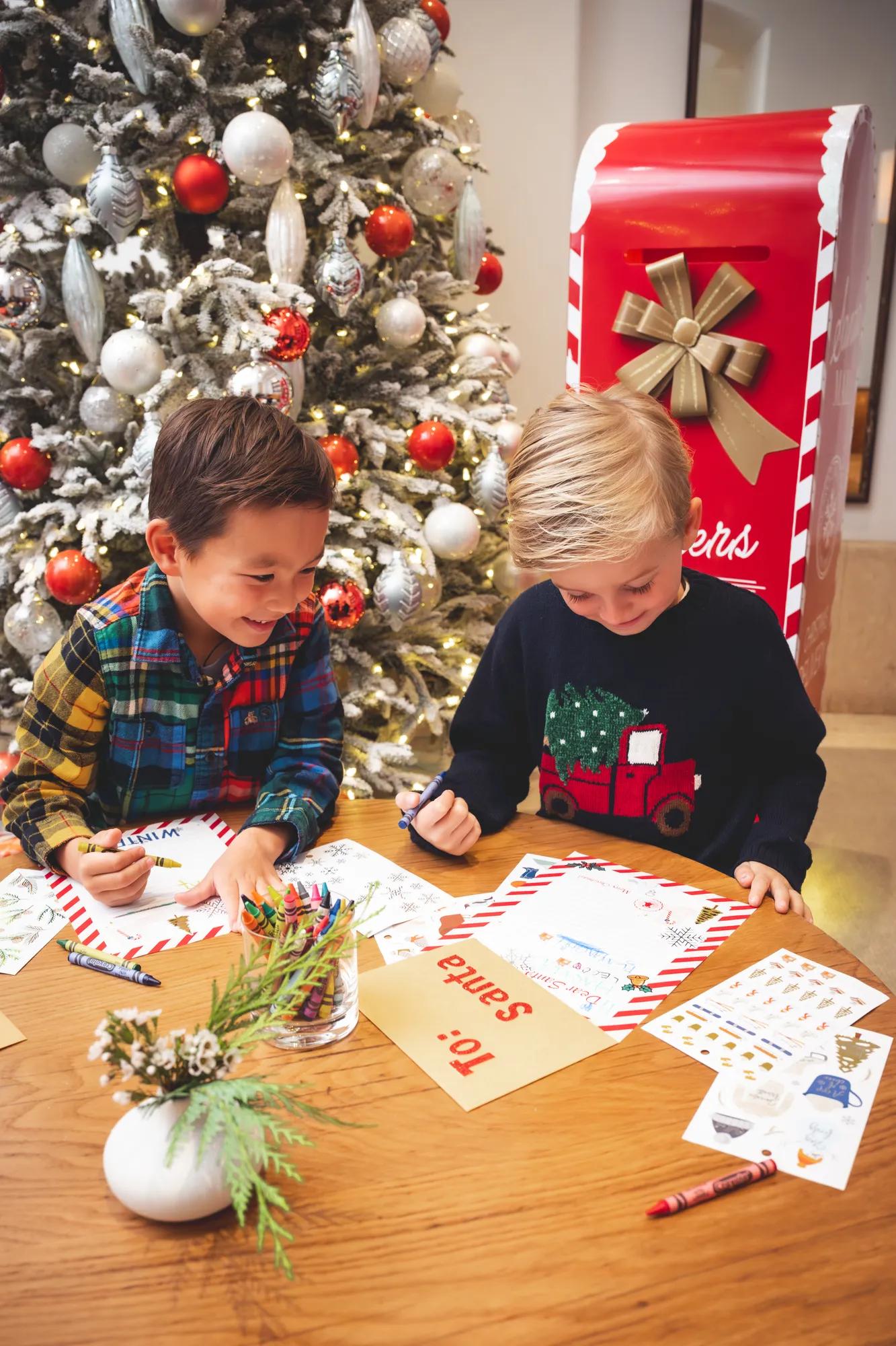 Children write holiday letters and color festive crafts at a table beside a decorated Christmas tree and oversized seasonal mailbox.