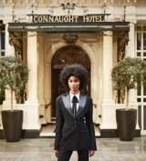 Woman in tailored black suit standing at entrance of Connaught Hotel, flanked by potted trees and ornate facade.