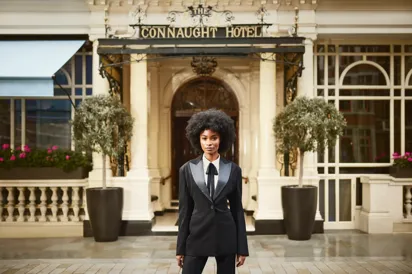 Woman in tailored black suit standing at entrance of Connaught Hotel, flanked by potted trees and ornate facade.