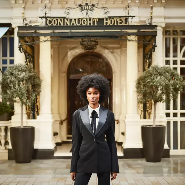Woman in tailored black suit standing at entrance of Connaught Hotel, flanked by potted trees and ornate facade.