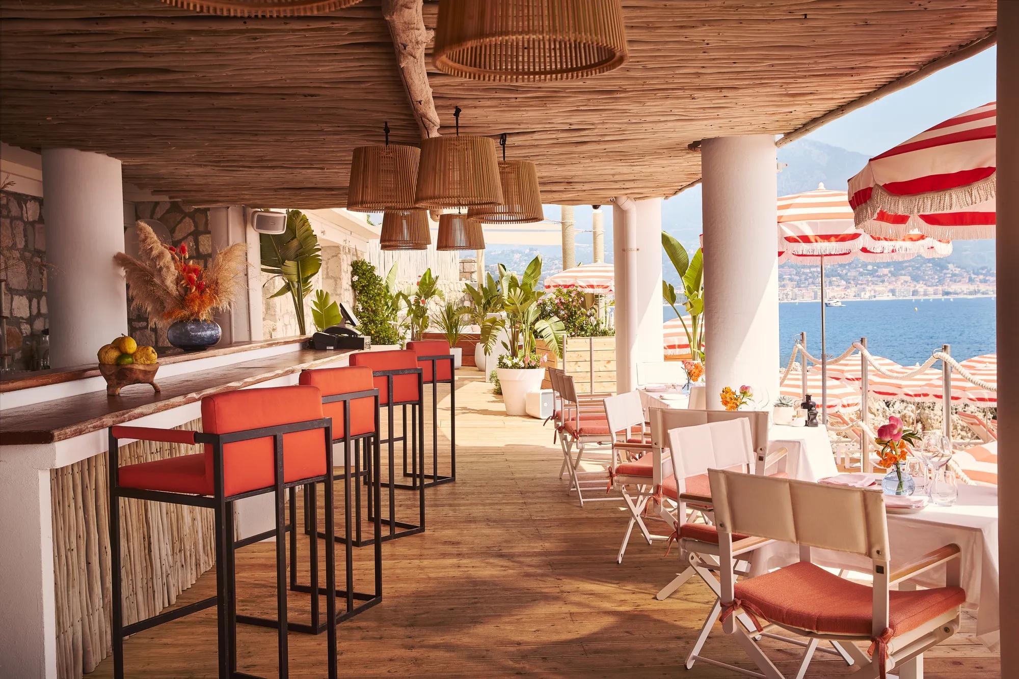 Terrasse ombragée avec tabourets corail, tables dressées et parasols rayés surplombant la mer
