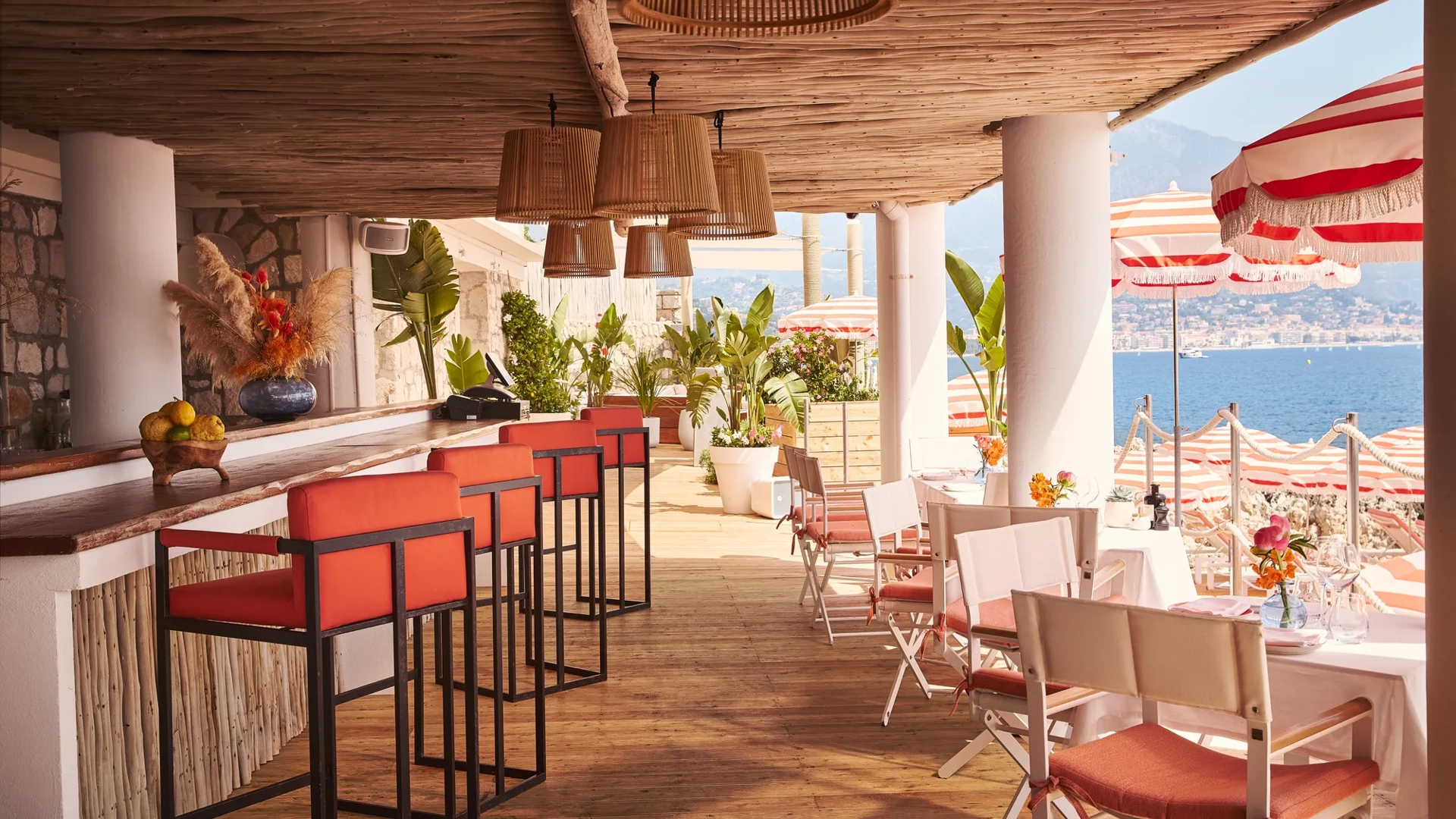 Terrasse ombragée avec tabourets corail, tables dressées et parasols rayés surplombant la mer