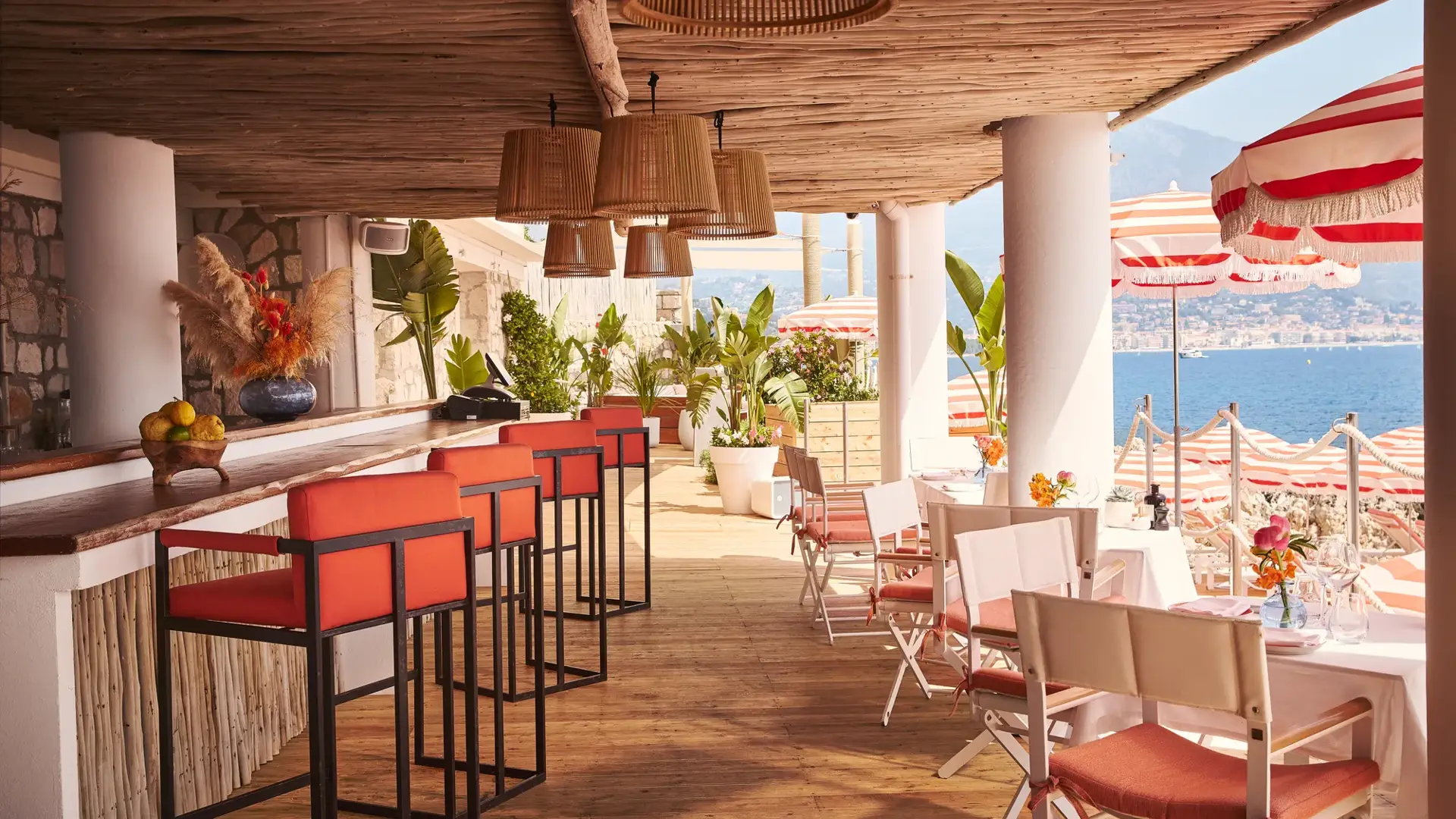 Terrasse ombragée avec tabourets corail, tables dressées et parasols rayés surplombant la mer