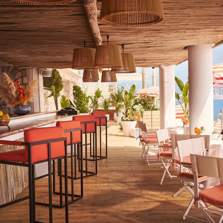 Terrasse ombragée avec tabourets corail, tables dressées et parasols rayés surplombant la mer
