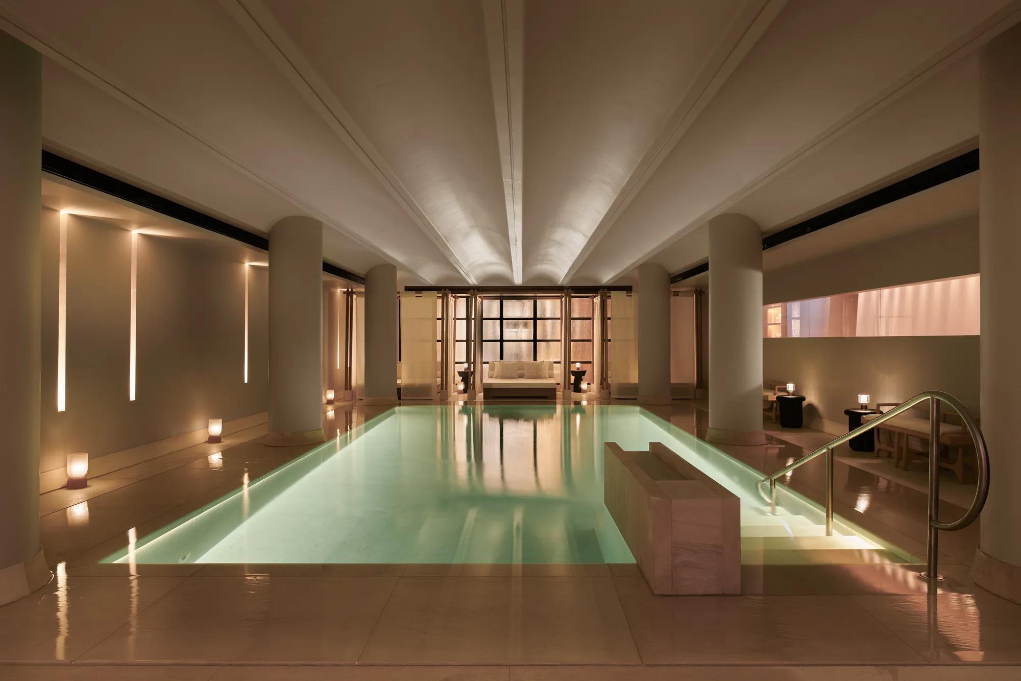 Serene indoor pool at The Maybourne Riviera, surrounded by soft lighting, smooth stone columns, and a seating area at the far end creating a tranquil, spa-like atmosphere.