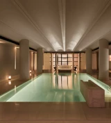 Serene indoor pool at The Maybourne Riviera, surrounded by soft lighting, smooth stone columns, and a seating area at the far end creating a tranquil, spa-like atmosphere.