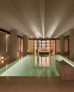 Serene indoor pool at The Maybourne Riviera, surrounded by soft lighting, smooth stone columns, and a seating area at the far end creating a tranquil, spa-like atmosphere.