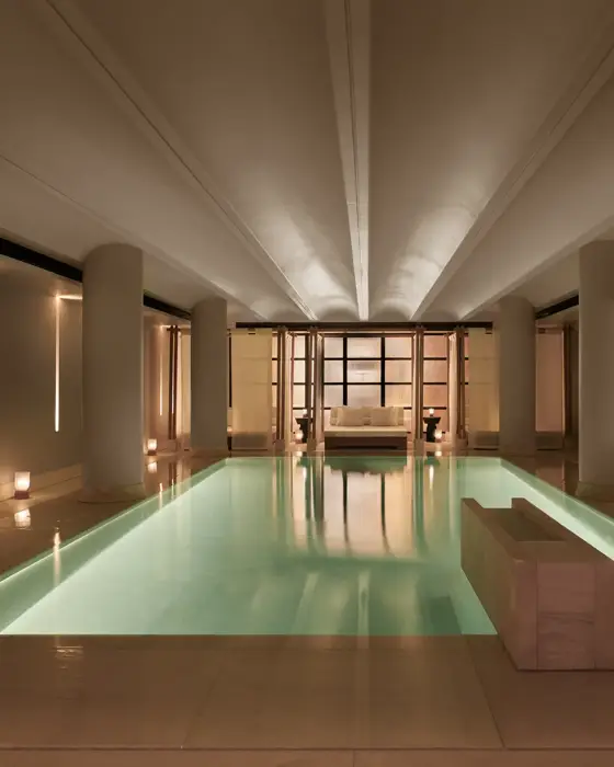 Serene indoor pool at The Maybourne Riviera, surrounded by soft lighting, smooth stone columns, and a seating area at the far end creating a tranquil, spa-like atmosphere.