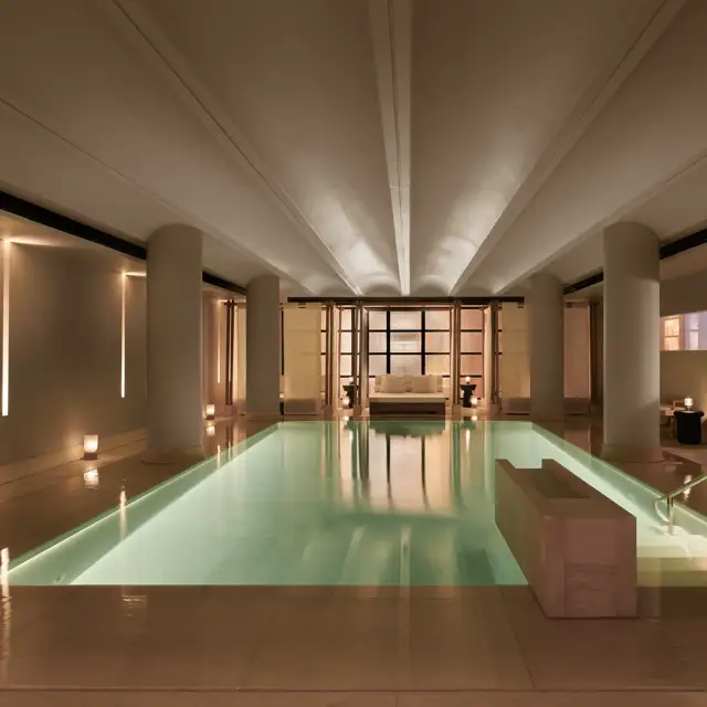 Serene indoor pool at The Maybourne Riviera, surrounded by soft lighting, smooth stone columns, and a seating area at the far end creating a tranquil, spa-like atmosphere.