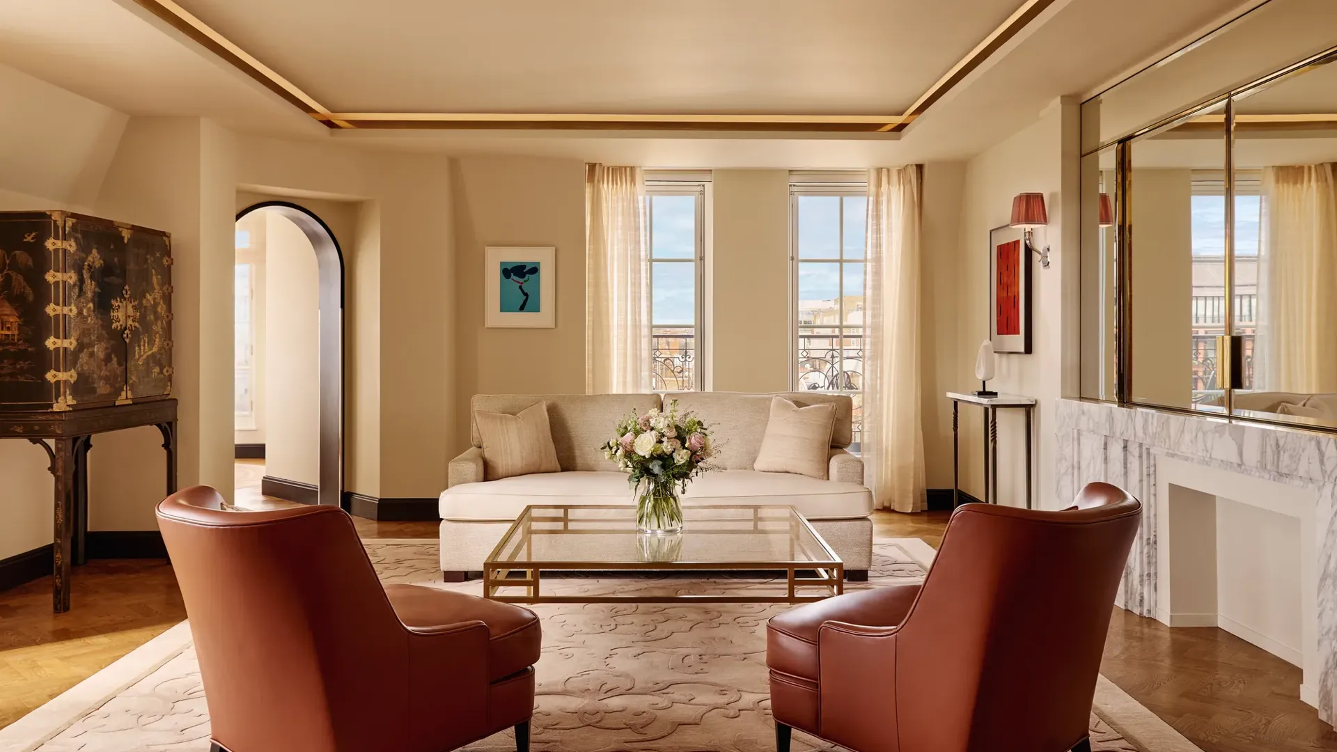 Bright living room with two brown leather armchairs facing a cream sofa, a glass coffee table with flowers, and large windows framed by sheer curtains, decorated in warm neutral tones with modern artwork.