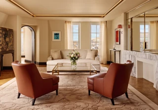 Bright living room with two brown leather armchairs facing a cream sofa, a glass coffee table with flowers, and large windows framed by sheer curtains, decorated in warm neutral tones with modern artwork.