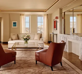 Bright living room with two brown leather armchairs facing a cream sofa, a glass coffee table with flowers, and large windows framed by sheer curtains, decorated in warm neutral tones with modern artwork.