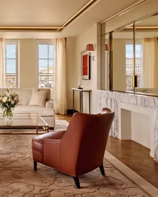 Bright living room with two brown leather armchairs facing a cream sofa, a glass coffee table with flowers, and large windows framed by sheer curtains, decorated in warm neutral tones with modern artwork.