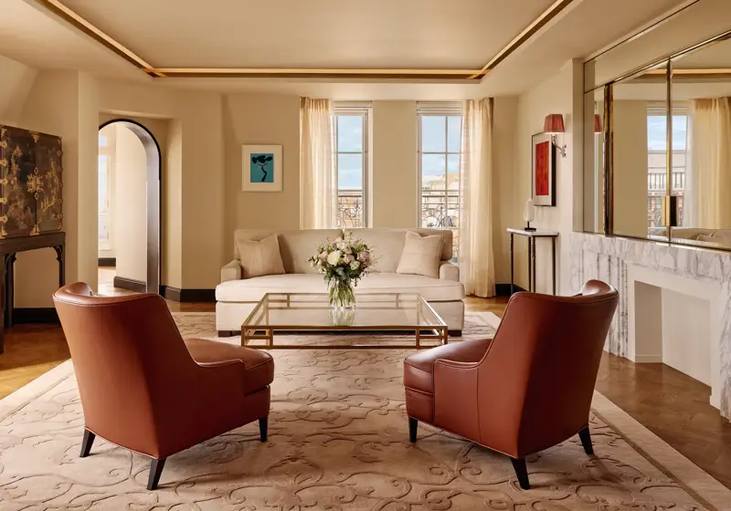 Bright living room with two brown leather armchairs facing a cream sofa, a glass coffee table with flowers, and large windows framed by sheer curtains, decorated in warm neutral tones with modern artwork.