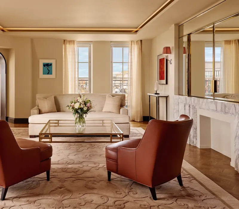Bright living room with two brown leather armchairs facing a cream sofa, a glass coffee table with flowers, and large windows framed by sheer curtains, decorated in warm neutral tones with modern artwork.