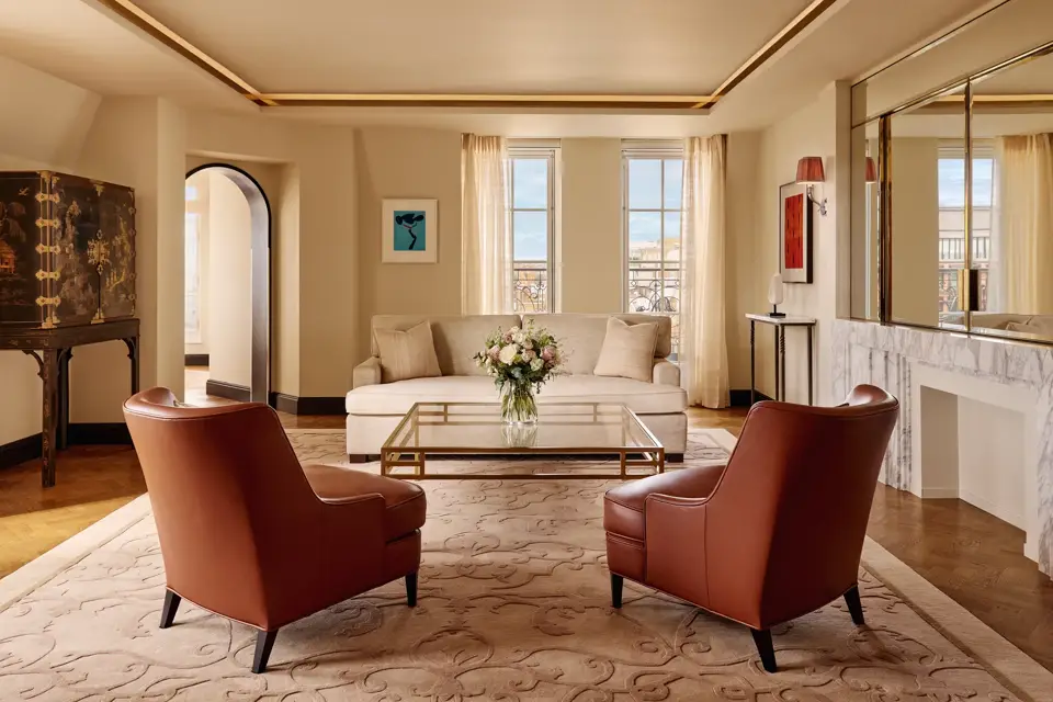 Bright living room with two brown leather armchairs facing a cream sofa, a glass coffee table with flowers, and large windows framed by sheer curtains, decorated in warm neutral tones with modern artwork.