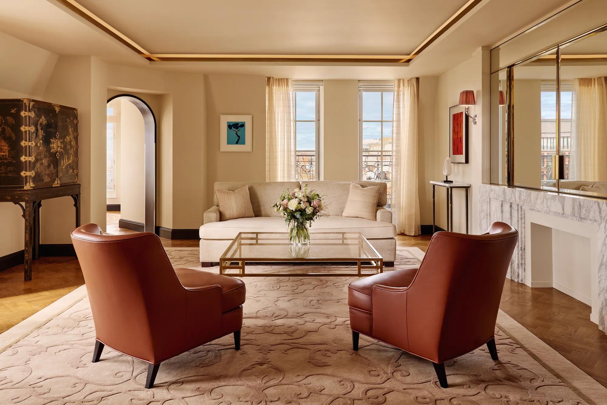 Bright living room with two brown leather armchairs facing a cream sofa, a glass coffee table with flowers, and large windows framed by sheer curtains, decorated in warm neutral tones with modern artwork.