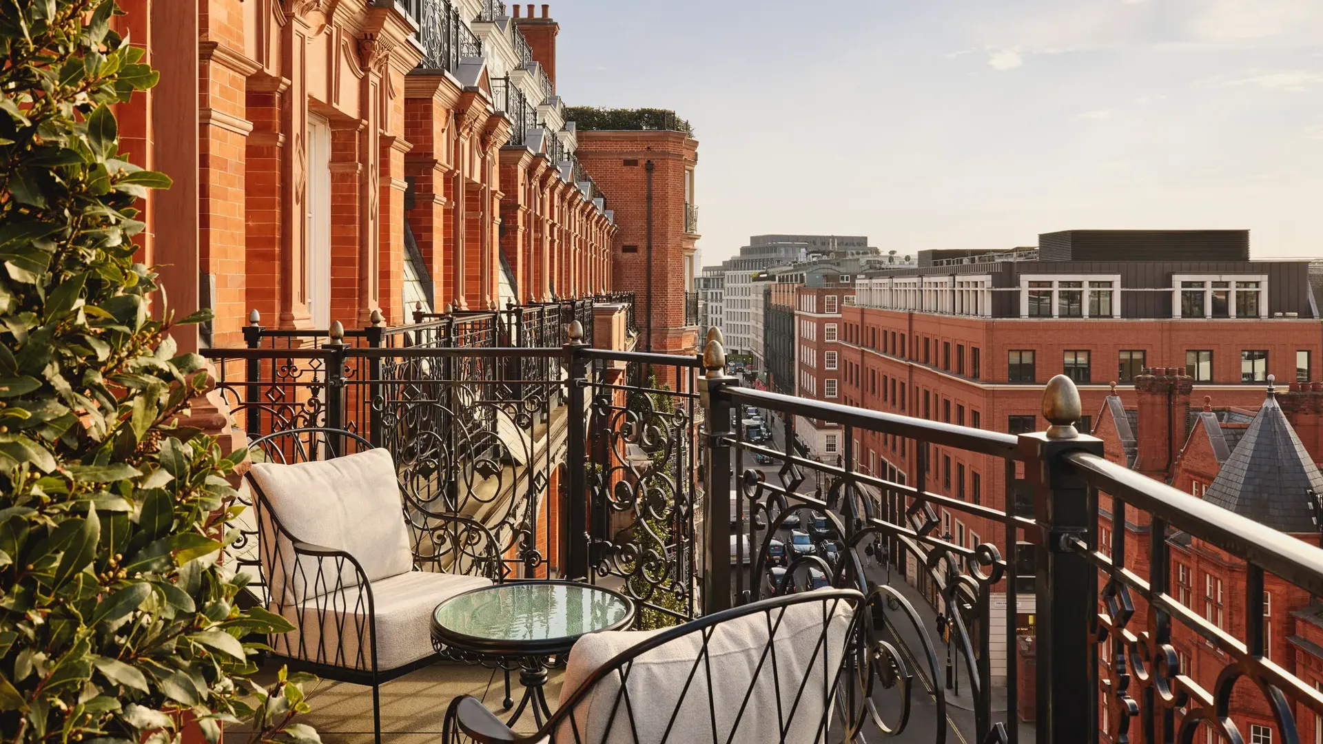 Balcony with ornate black wrought-iron railings and cushioned chairs around a glass-topped table, overlooking red-brick buildings and city rooftops under a clear sky.