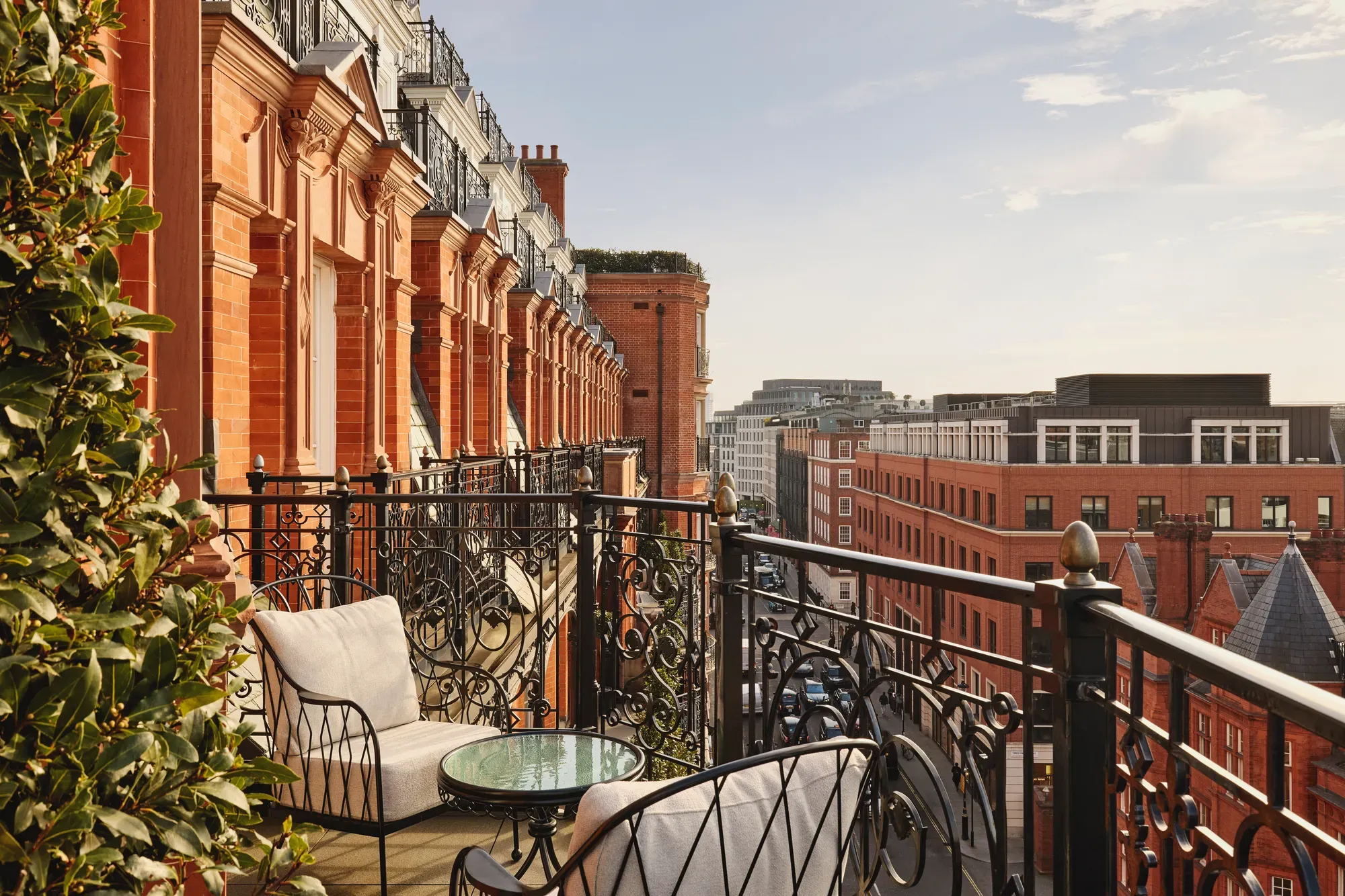 Balcony with ornate black wrought-iron railings and cushioned chairs around a glass-topped table, overlooking red-brick buildings and city rooftops under a clear sky.