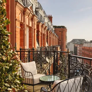 Balcony with ornate black wrought-iron railings and cushioned chairs around a glass-topped table, overlooking red-brick buildings and city rooftops under a clear sky.