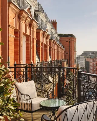 Balcony with ornate black wrought-iron railings and cushioned chairs around a glass-topped table, overlooking red-brick buildings and city rooftops under a clear sky.