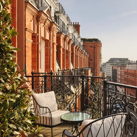 Balcony with ornate black wrought-iron railings and cushioned chairs around a glass-topped table, overlooking red-brick buildings and city rooftops under a clear sky.