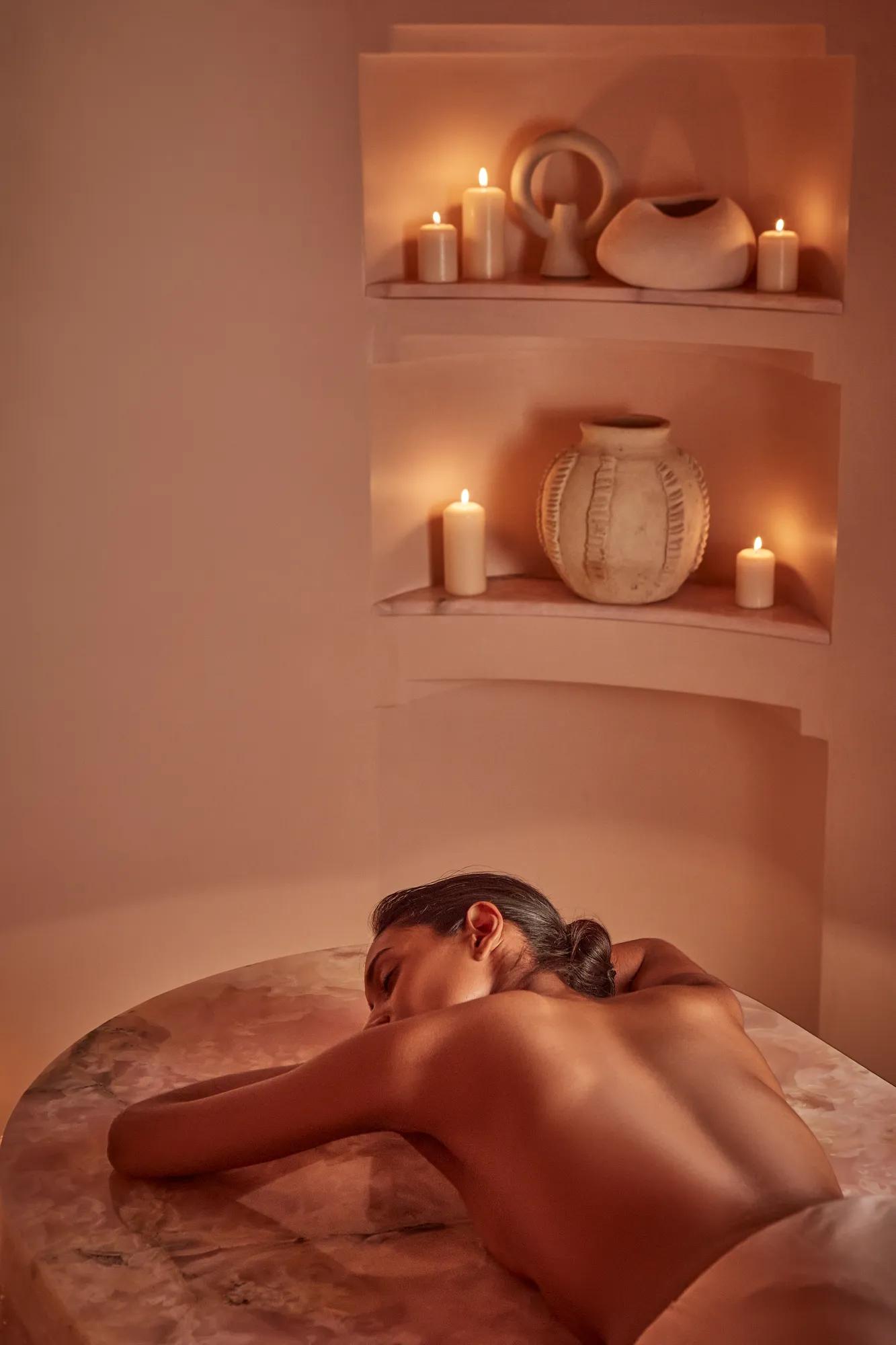 Woman lying face down on a massage table in a softly lit spa room with candles and ceramic decor in niches.