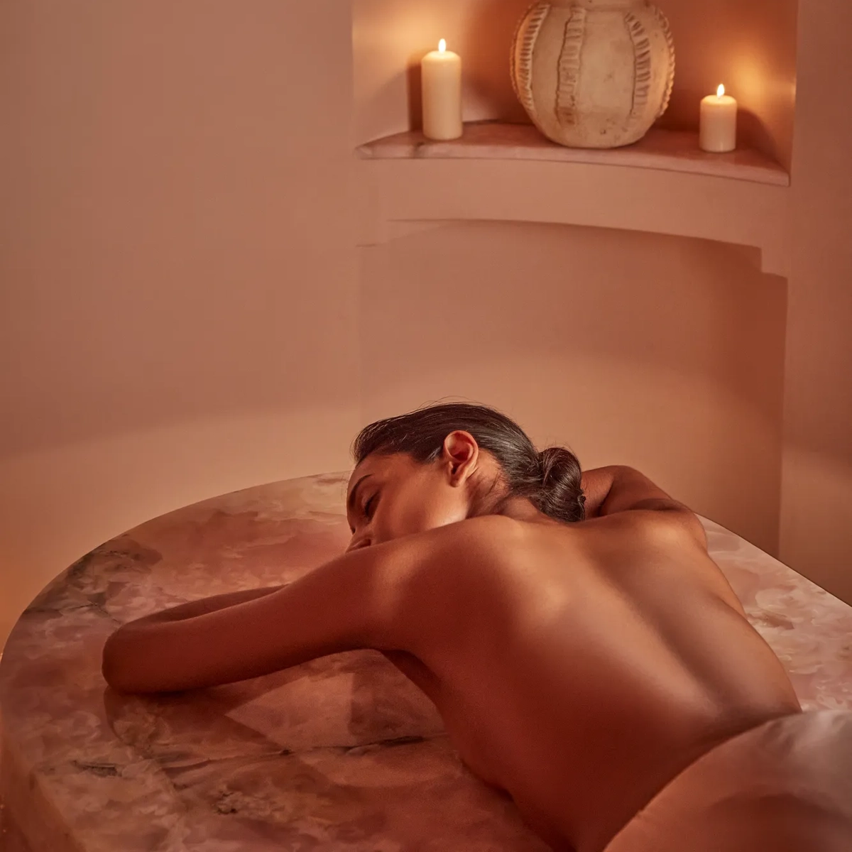 Woman lying face down on a massage table in a softly lit spa room with candles and ceramic decor in niches.