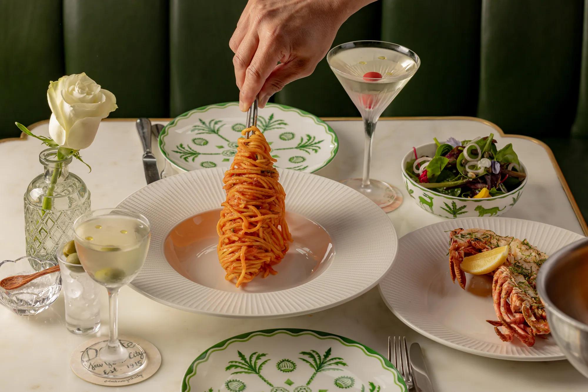 Hand lifting a forkful of tomato-coated spaghetti from a plate, with lobster, mixed salad and cocktails arranged on an elegant dining table.