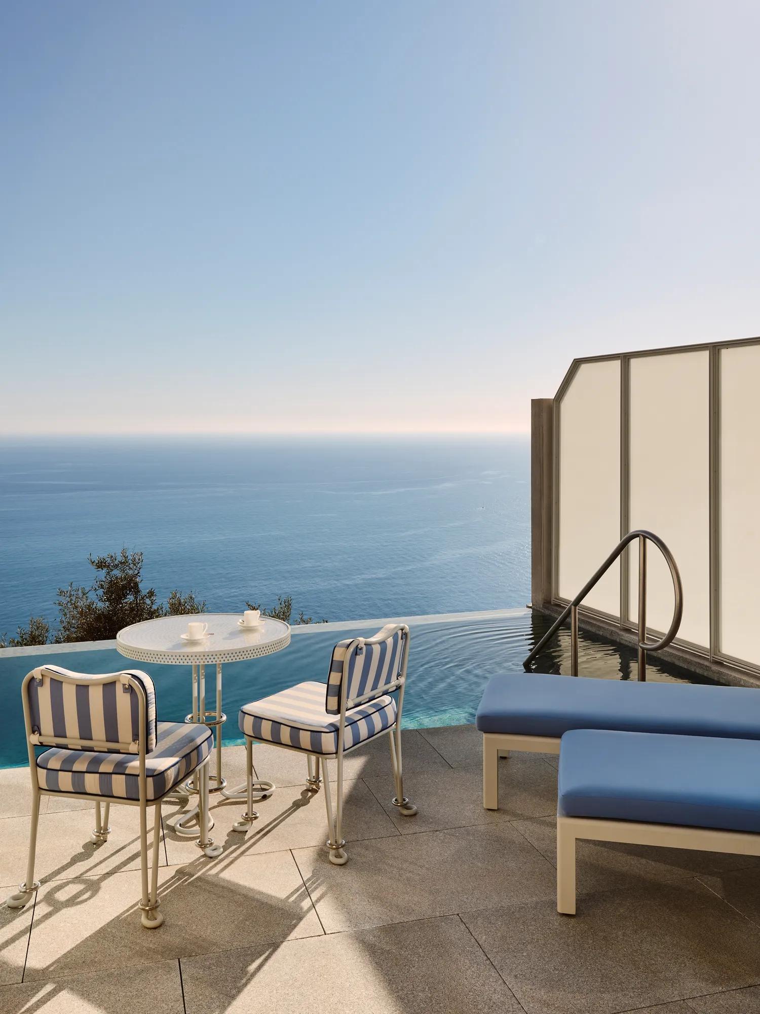Terrasse privée avec chaises rayées, petite table et transats au bord d’une piscine à débordement surplombant la mer Méditerranée.