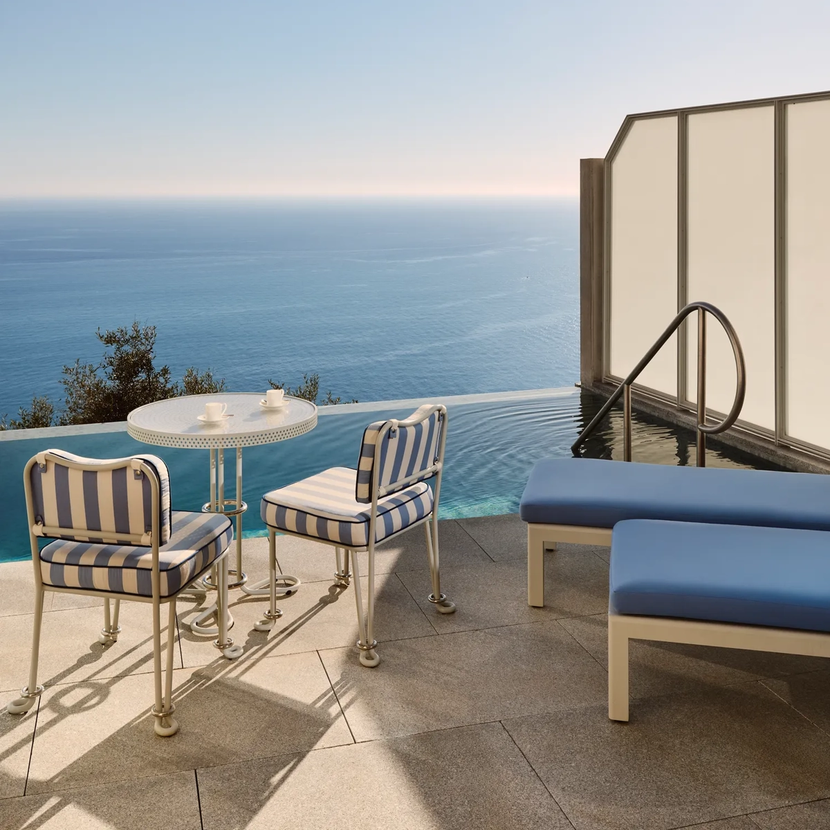 Terrasse privée avec chaises rayées, petite table et transats au bord d’une piscine à débordement surplombant la mer Méditerranée.