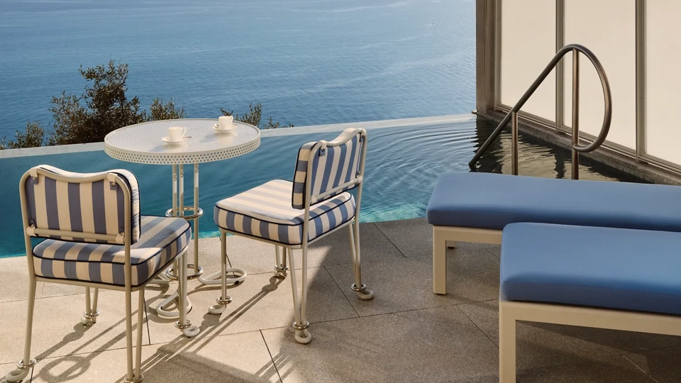 Terrasse privée avec chaises rayées, petite table et transats au bord d’une piscine à débordement surplombant la mer Méditerranée.