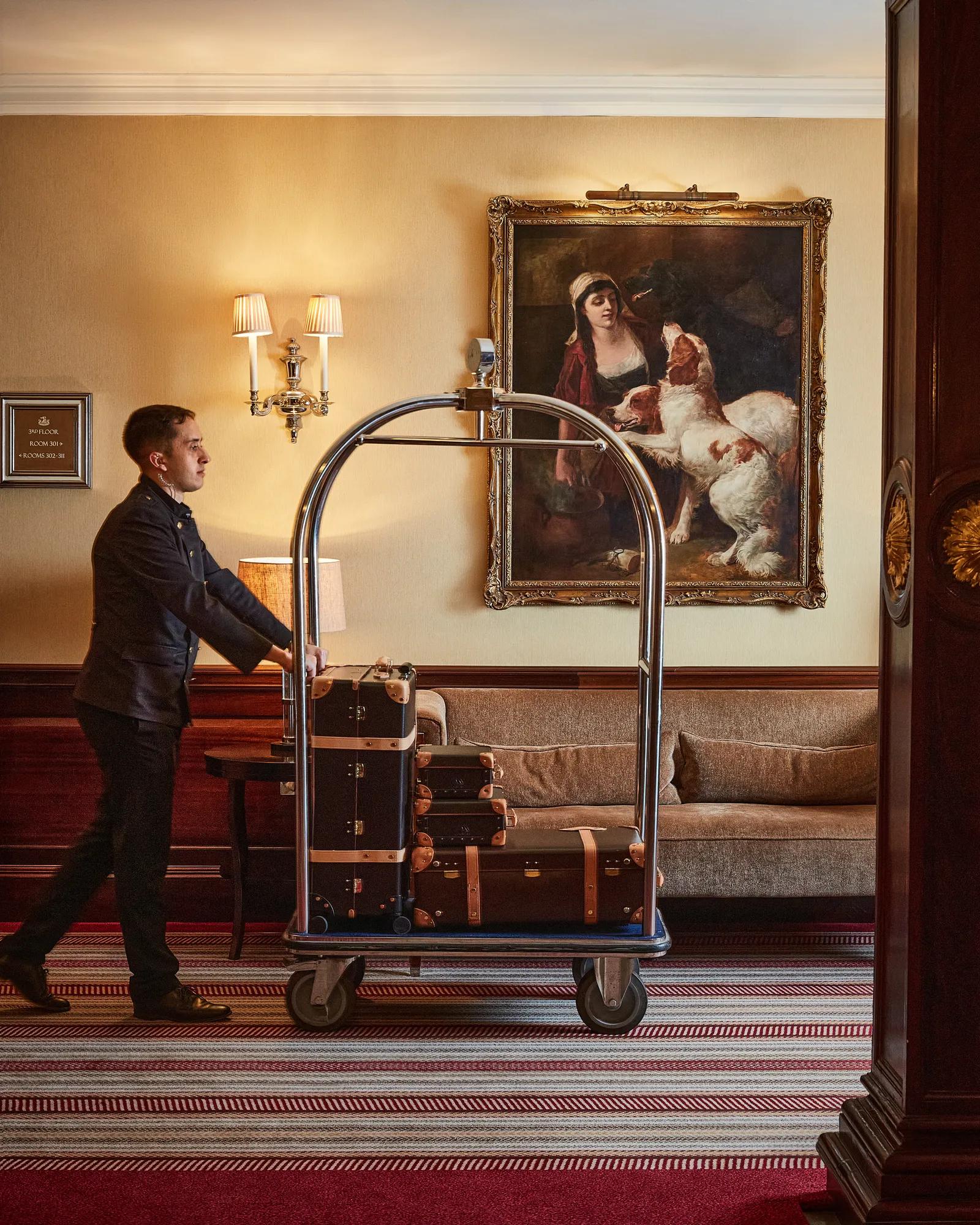 Hotel porter pushes a luggage cart stacked with suitcases through a lounge with a sofa and framed painting.