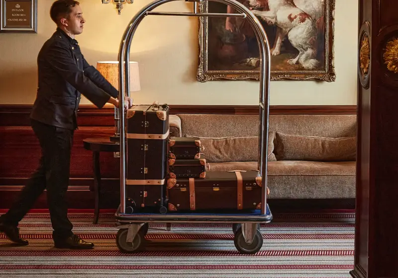 Hotel porter pushes a luggage cart stacked with suitcases through a lounge with a sofa and framed painting.