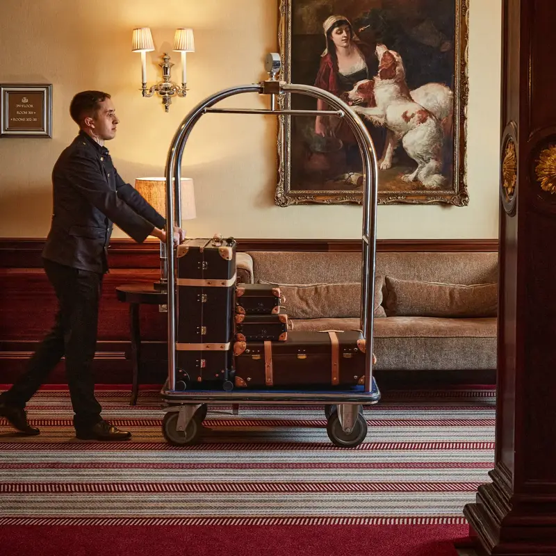 Hotel porter pushes a luggage cart stacked with suitcases through a lounge with a sofa and framed painting.