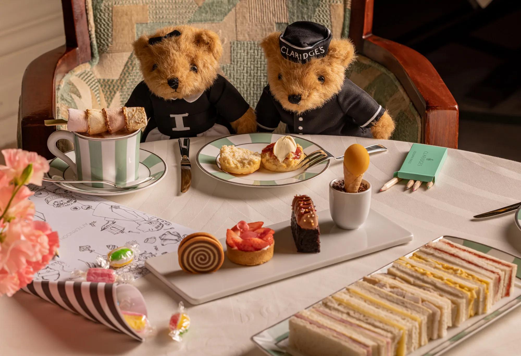 Two teddy bears in Claridge’s uniforms enjoy afternoon tea with cakes, sandwiches and crayons on a white table.