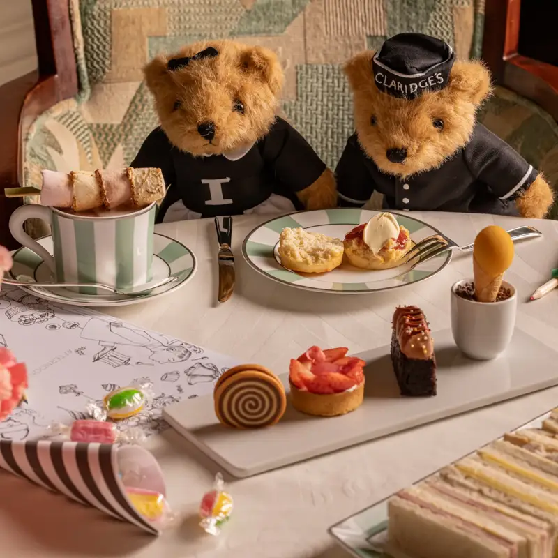 Two teddy bears in Claridge’s uniforms enjoy afternoon tea with cakes, sandwiches and crayons on a white table.