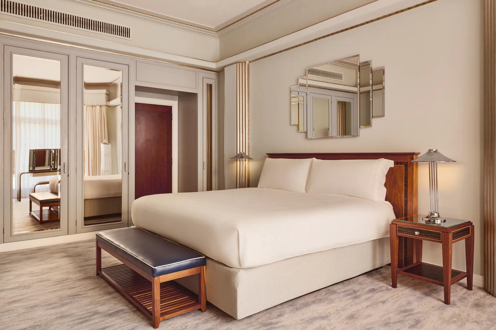 Elegant bedroom with a large bed framed by Art Deco–style mirrored panels and wooden furniture, including a leather bench and bedside tables, softly lit by wall lights and natural daylight.