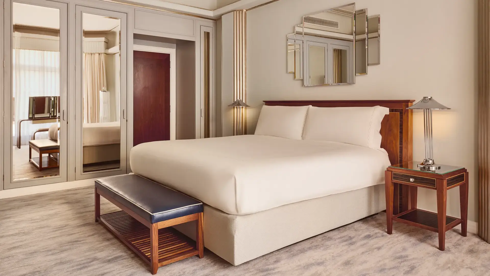 Elegant bedroom with a large bed framed by Art Deco–style mirrored panels and wooden furniture, including a leather bench and bedside tables, softly lit by wall lights and natural daylight.