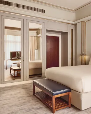 Elegant bedroom with a large bed framed by Art Deco–style mirrored panels and wooden furniture, including a leather bench and bedside tables, softly lit by wall lights and natural daylight.
