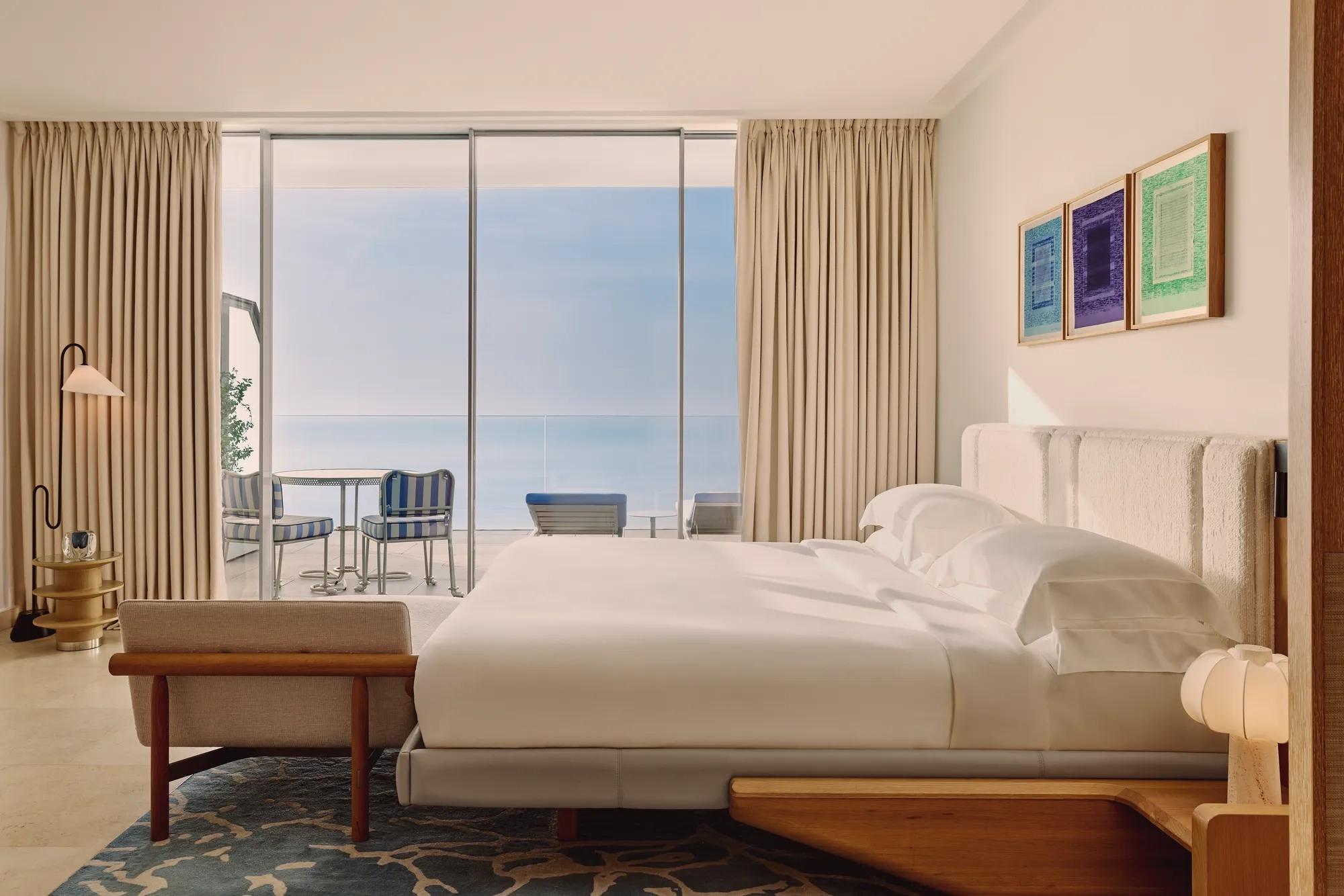 Hotel bedroom with large bed, modern furnishings, and glass doors opening to a terrace with striped chairs and sea views.