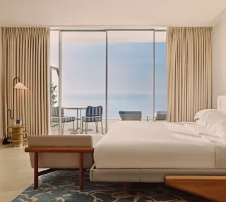 Hotel bedroom with large bed, modern furnishings, and glass doors opening to a terrace with striped chairs and sea views.
