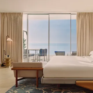 Hotel bedroom with large bed, modern furnishings, and glass doors opening to a terrace with striped chairs and sea views.