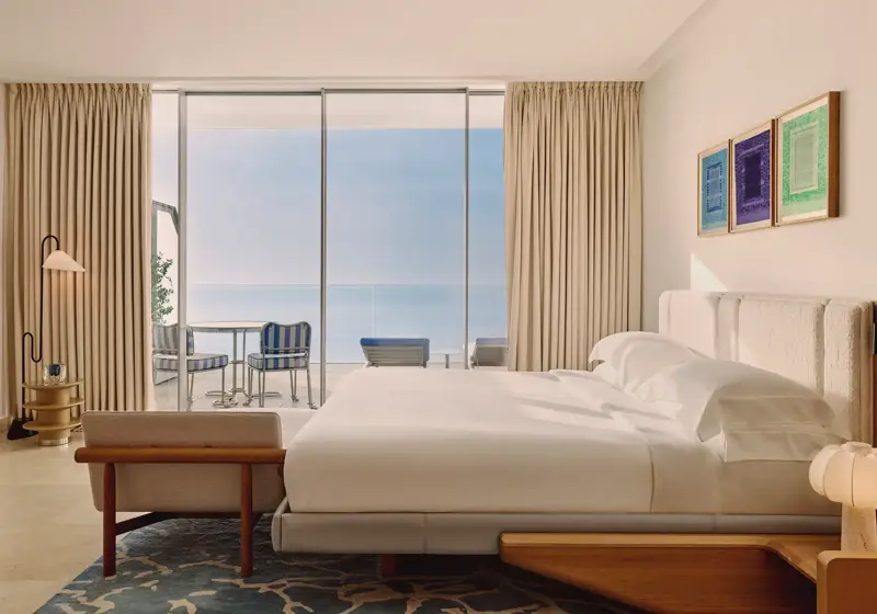 Hotel bedroom with large bed, modern furnishings, and glass doors opening to a terrace with striped chairs and sea views.