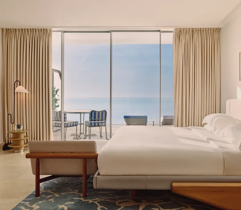 Hotel bedroom with large bed, modern furnishings, and glass doors opening to a terrace with striped chairs and sea views.