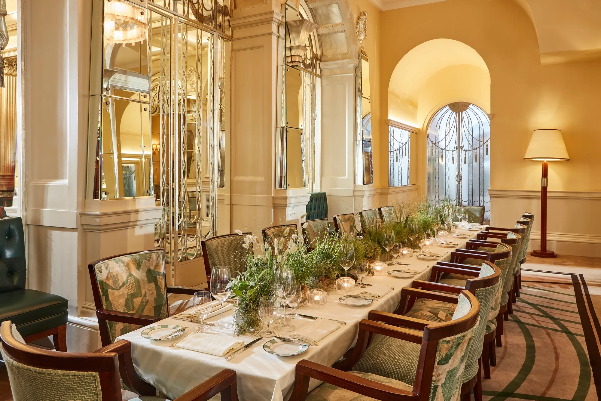 Elegant private dining table set for an event in a mirrored Art Deco room with soft yellow walls, green upholstered chairs, candlelight, and a long centrepiece of fresh greenery.