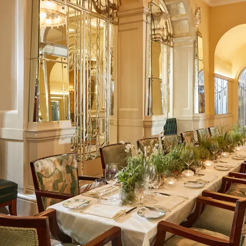 Elegant private dining table set for an event in a mirrored Art Deco room with soft yellow walls, green upholstered chairs, candlelight, and a long centrepiece of fresh greenery.
