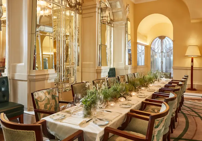 Elegant private dining table set for an event in a mirrored Art Deco room with soft yellow walls, green upholstered chairs, candlelight, and a long centrepiece of fresh greenery.