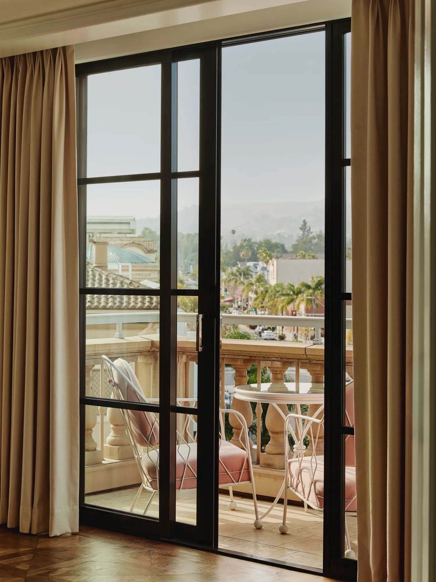 Balcony scene framed by black‑steel doors: pink‑cushioned chairs and round table behind a stone balustrade, overlooking palm‑lined Beverly Hills streets.