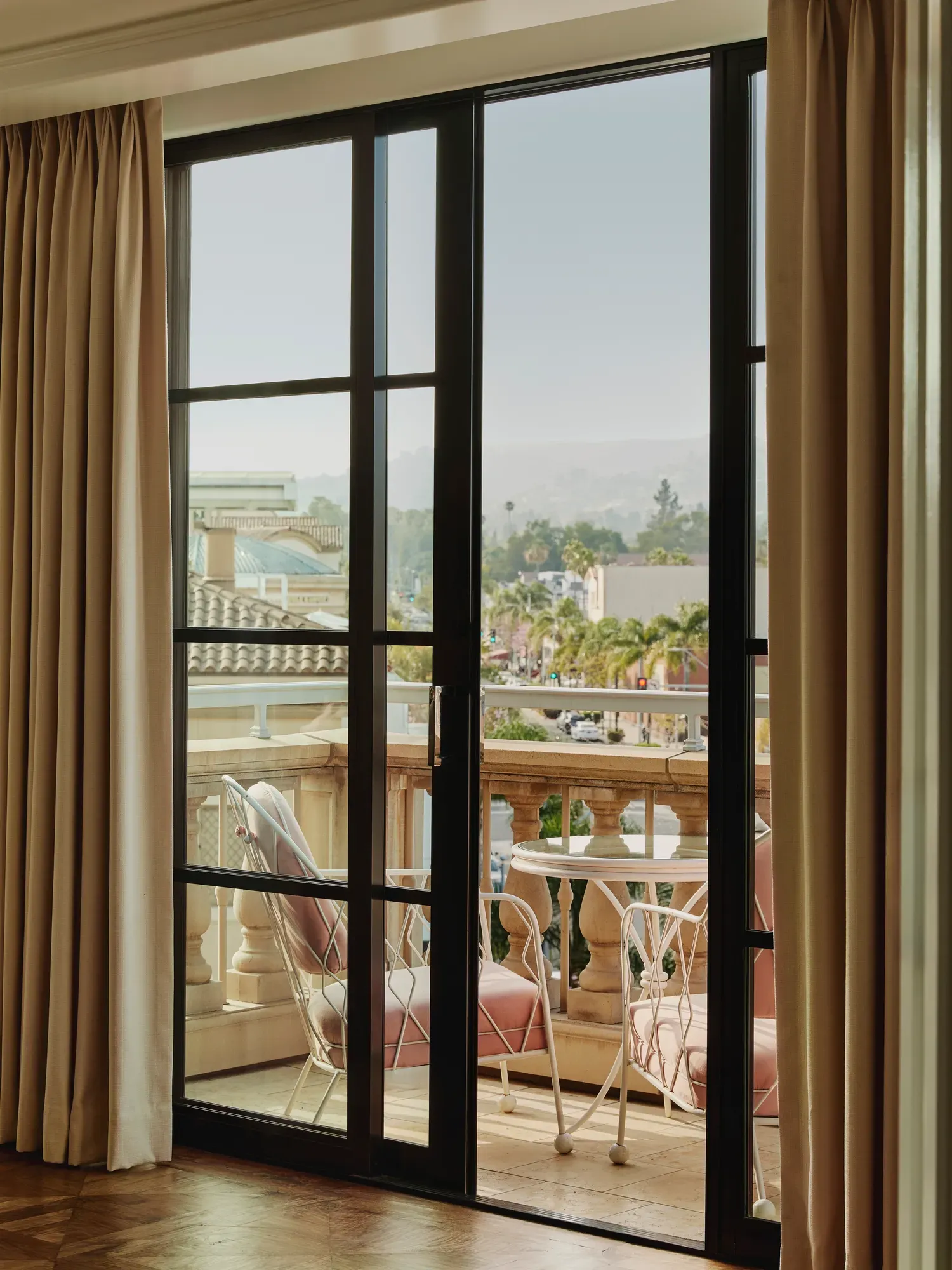 Balcony scene framed by black‑steel doors: pink‑cushioned chairs and round table behind a stone balustrade, overlooking palm‑lined Beverly Hills streets.